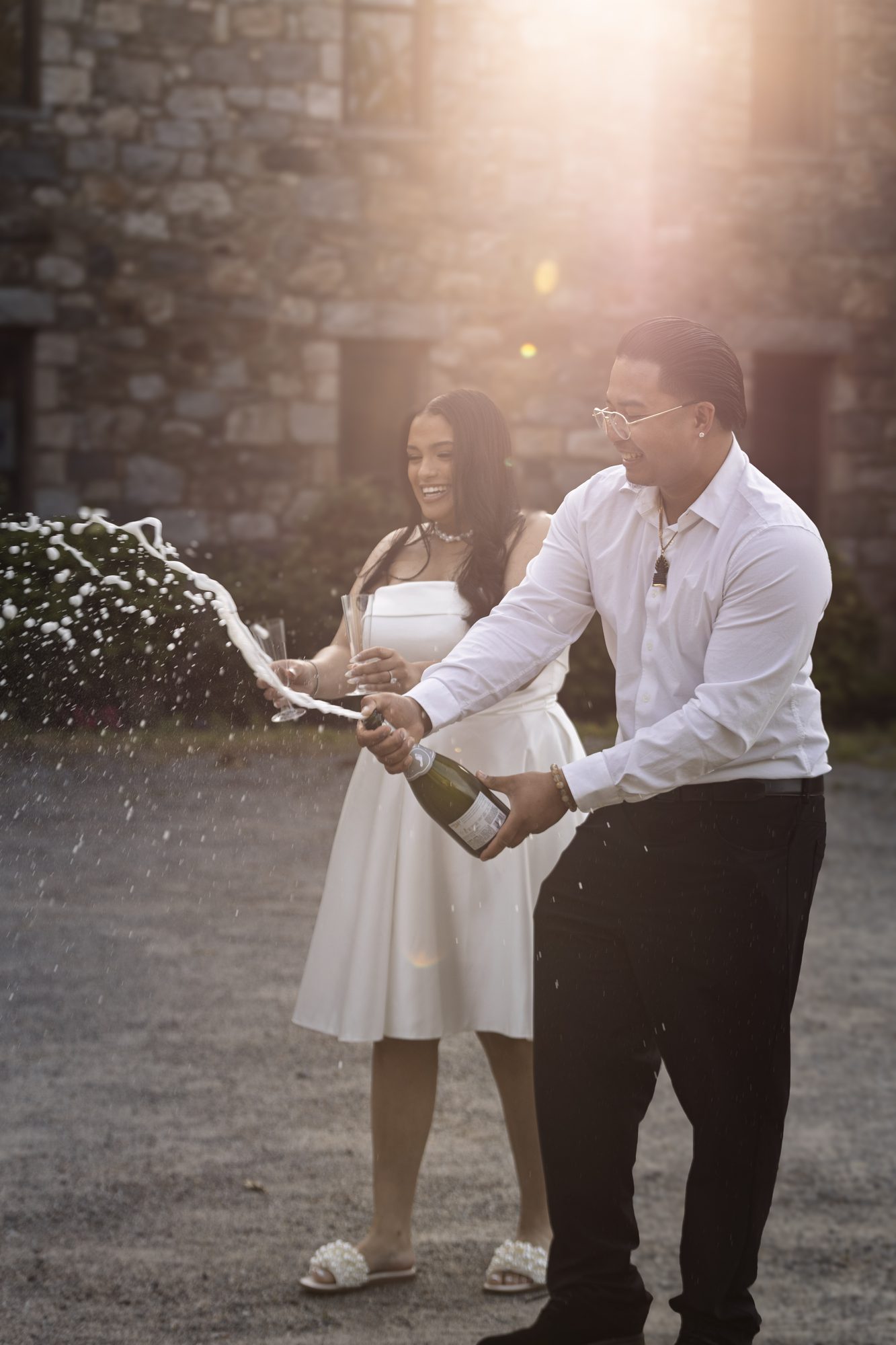 Man spraying champagne beside a smiling woman in a white dress, sunlight flaring behind a stone building