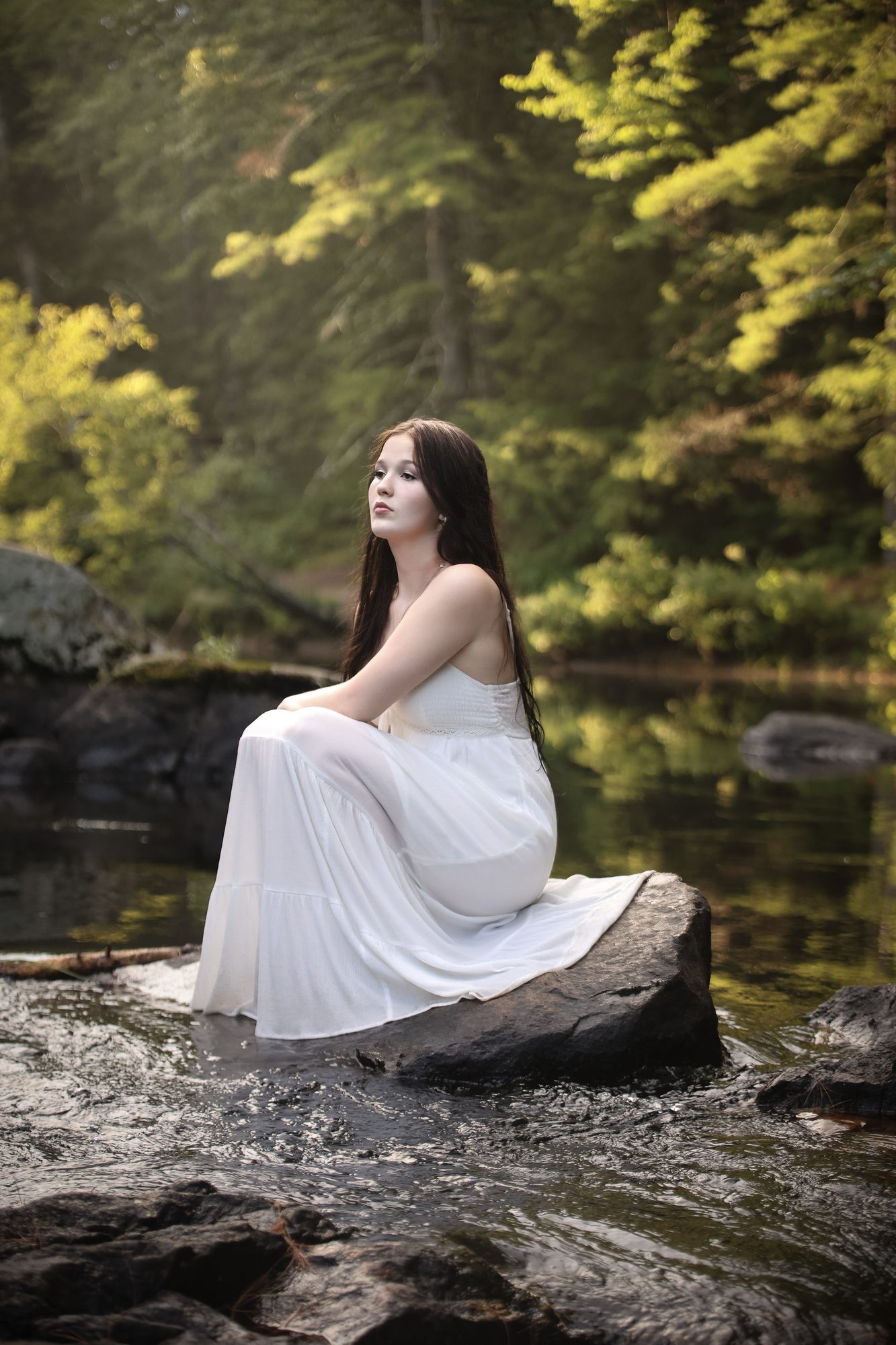 Young woman in a flowing white dress seated on a rock in a forest stream