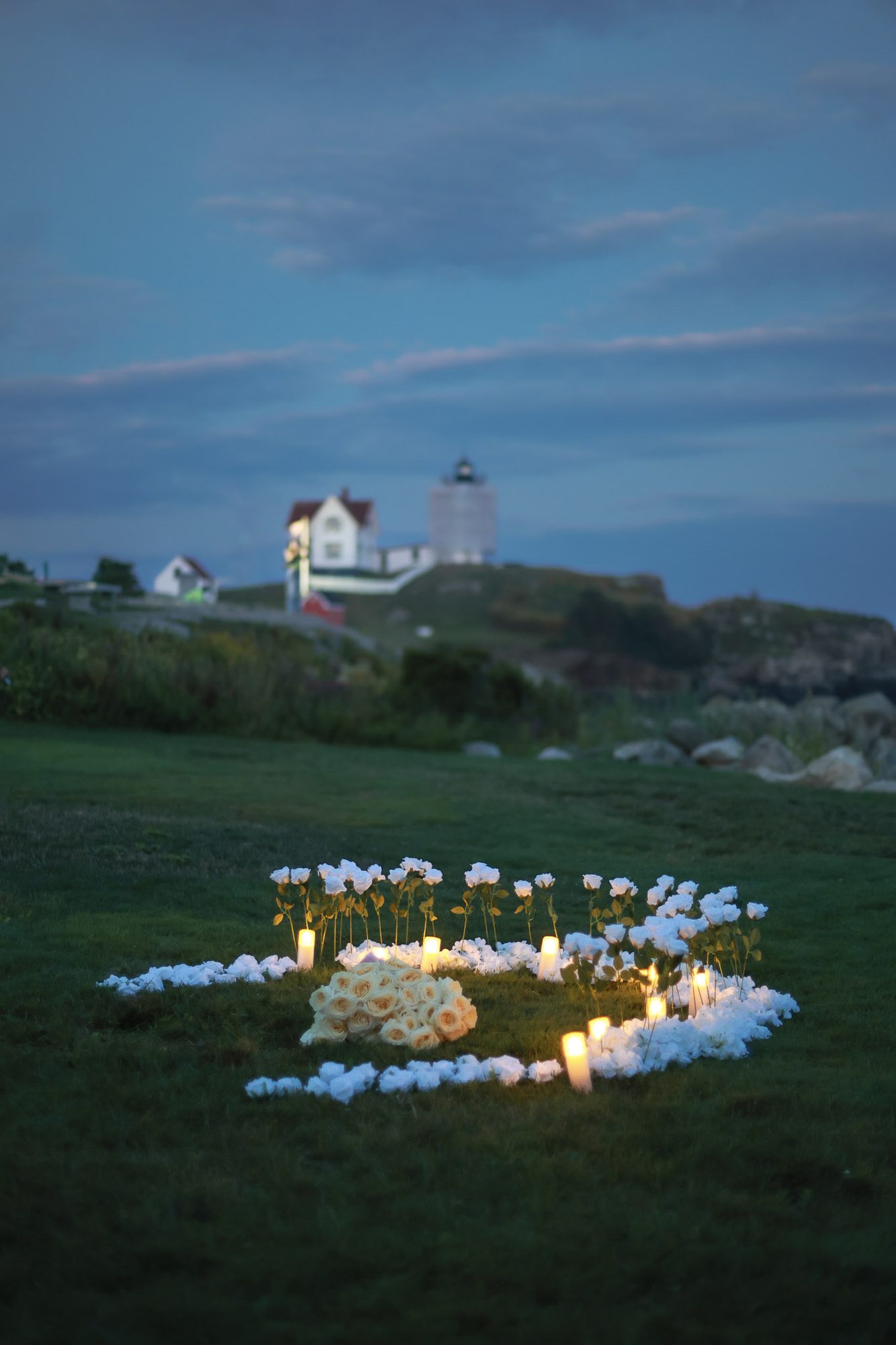 Ring of white roses and glowing candles set on the grass for a proposal, with a lighthouse on the rocky point beyond at dusk