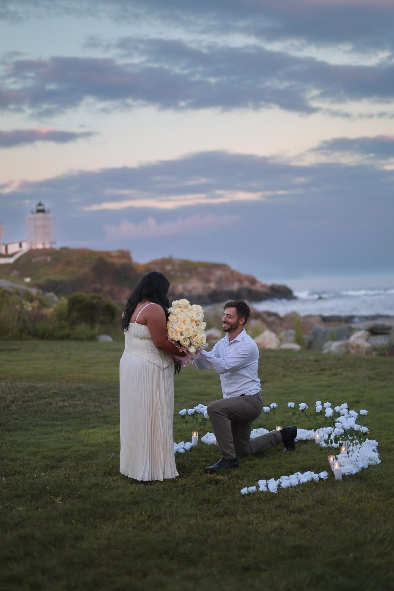 Man on one knee proposing near a lighthouse at dusk, inside a ring of white roses
