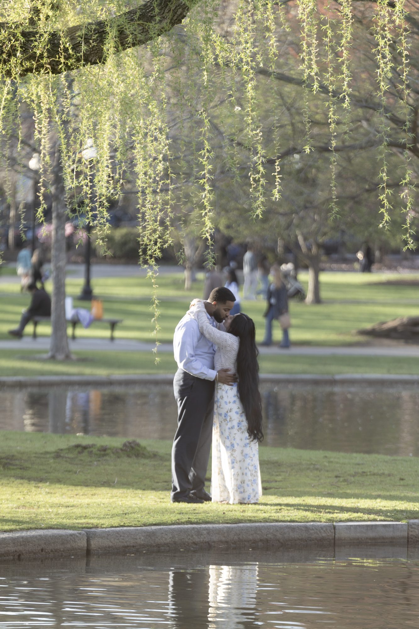 Couple embracing beside a park pond beneath a weeping willow, their reflection in the water
