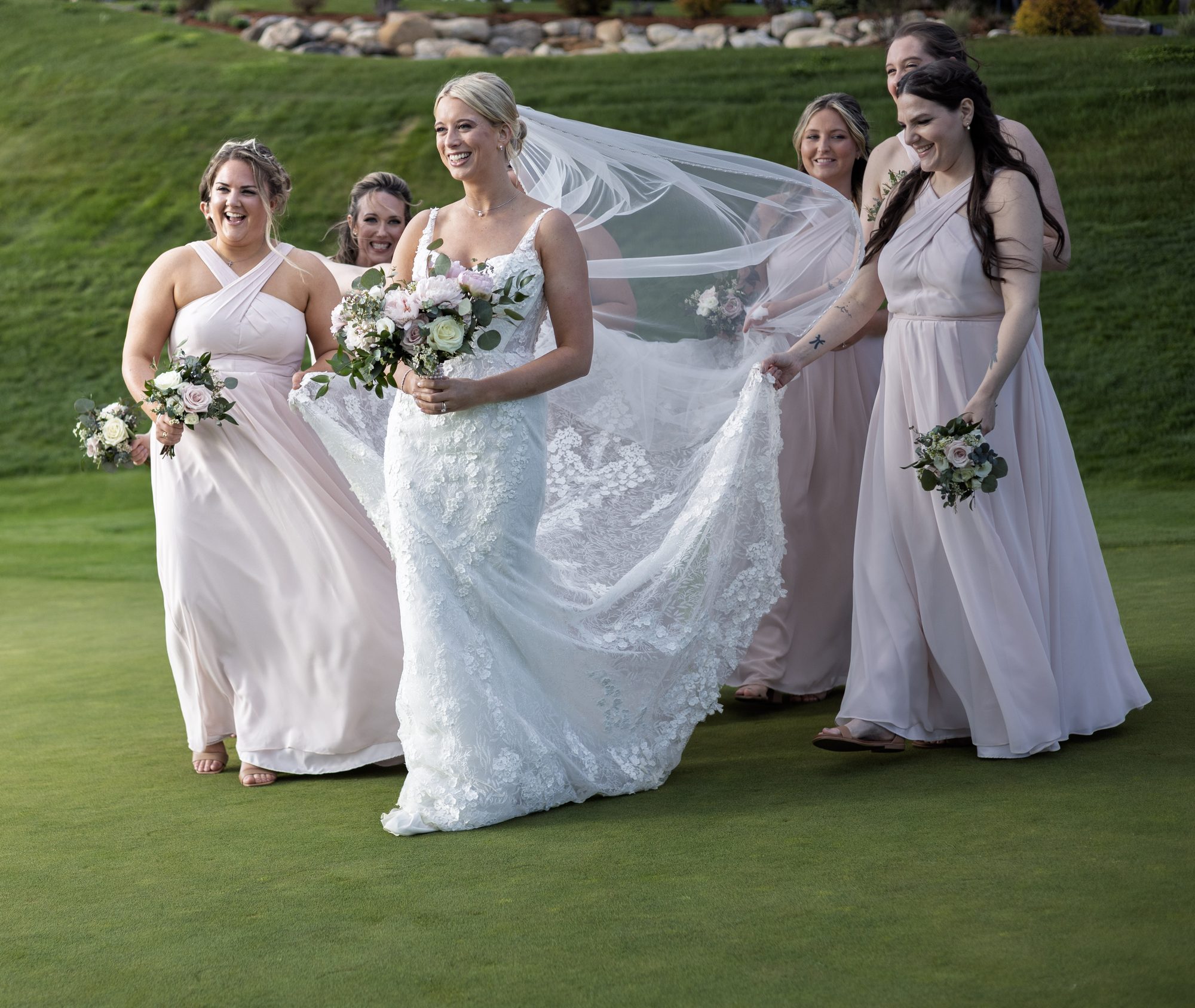 Bride walking across the grass with her bridesmaids in blush gowns, her veil catching the breeze