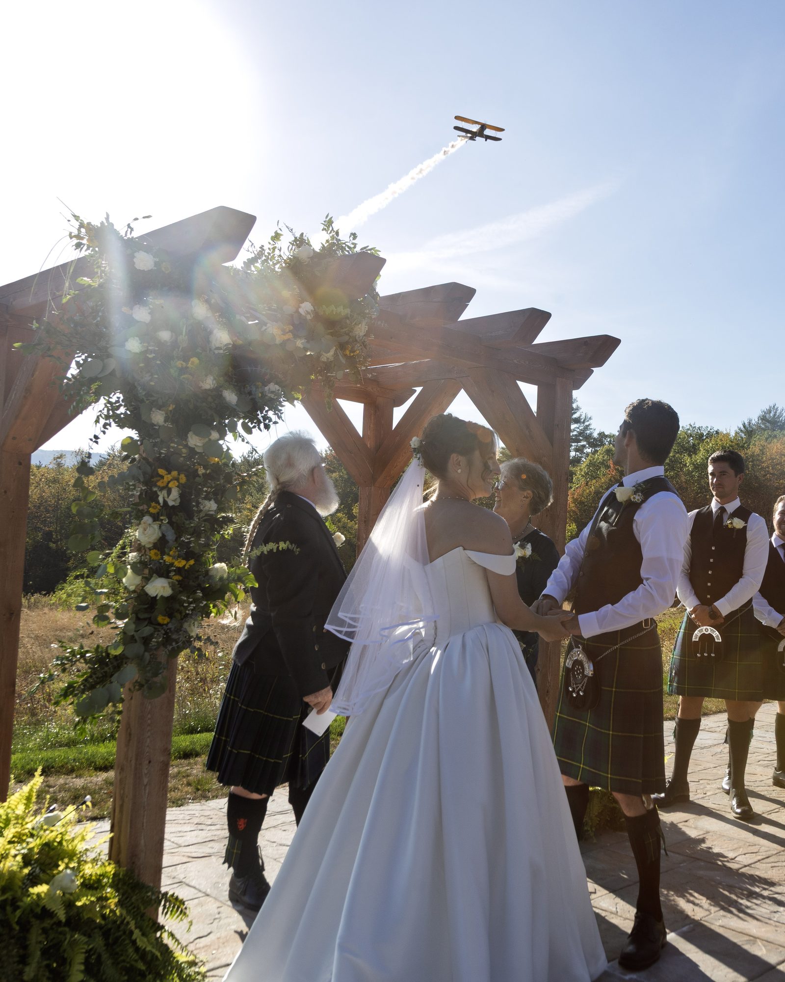 Bride and groom holding hands under a flower-draped wooden arch during an outdoor ceremony, groomsmen in kilts looking on as a biplane flies overhead