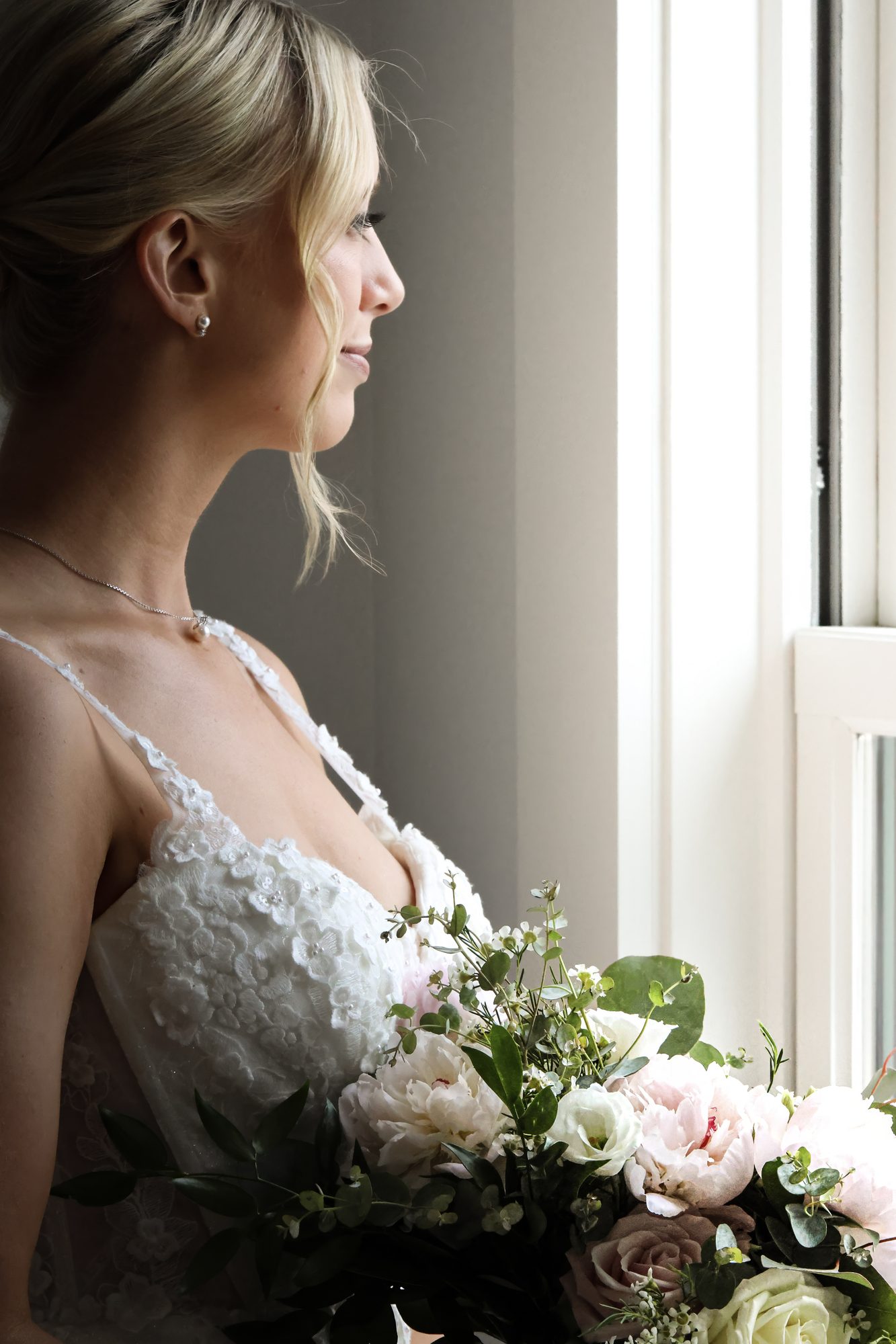 Bride in profile by a sunlit window, holding a bouquet of blush roses and greenery