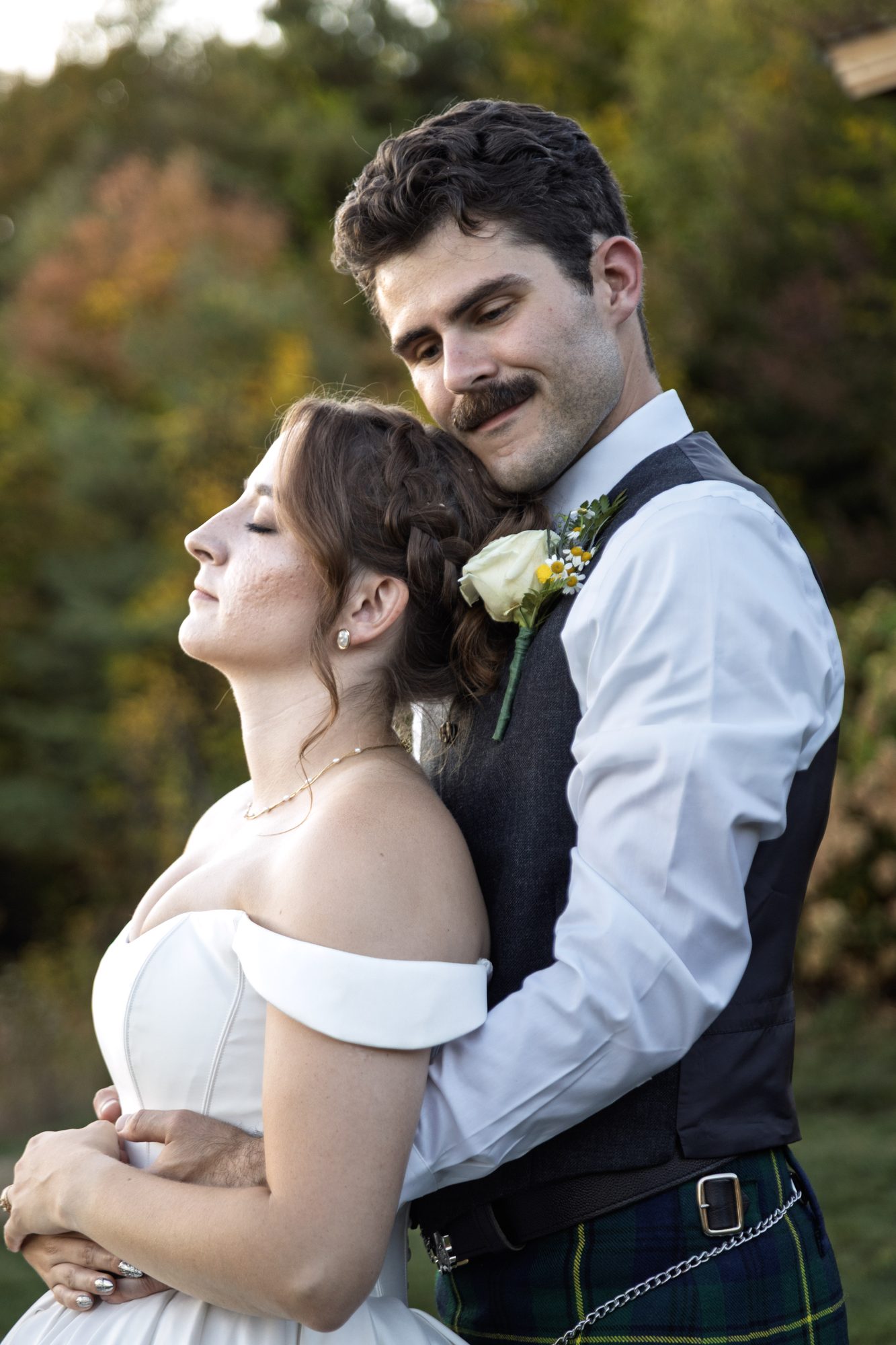 Groom in a kilt holding the bride from behind as she leans back with her eyes closed, autumn trees behind them